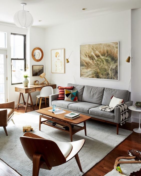 A small mid-century modern living room with a gray sofa, a tiered coffee table, a leather armchair and a neutral chair, a desk and a white chair, some art