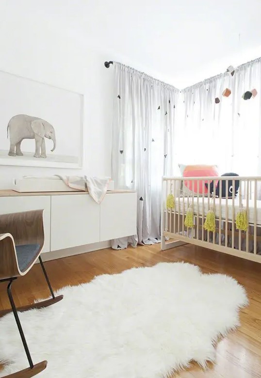 a small neutral Scandi nursery with a stained crib, white floating dresser, rocking chair, rug, artwork and some sheer curtains