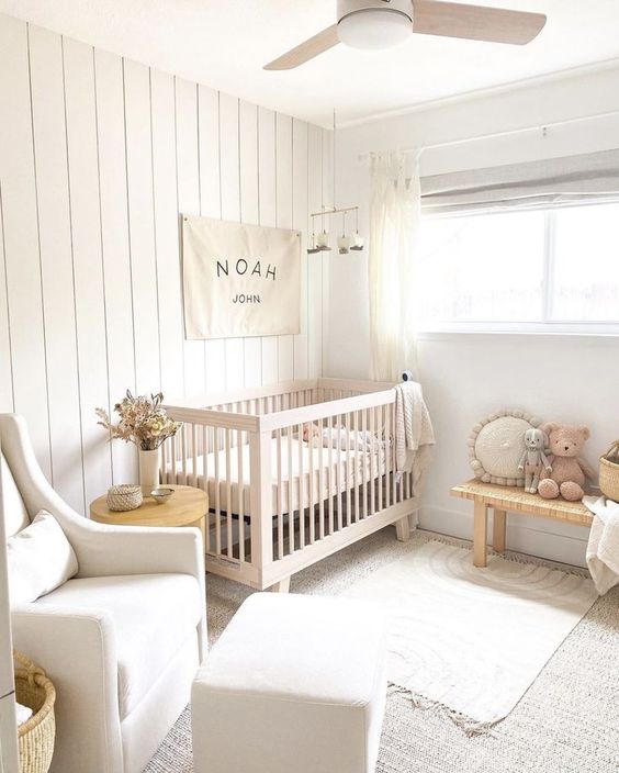 a small neutral nursery with a stained crib, woven bench, neutral chair, ottoman, layered rugs and pretty decor