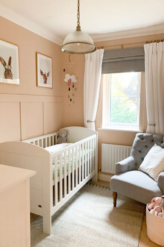 a small pastel-colored nursery with pink walls, a white crib, a gray chair, a gallery wall, some decor and layered curtains