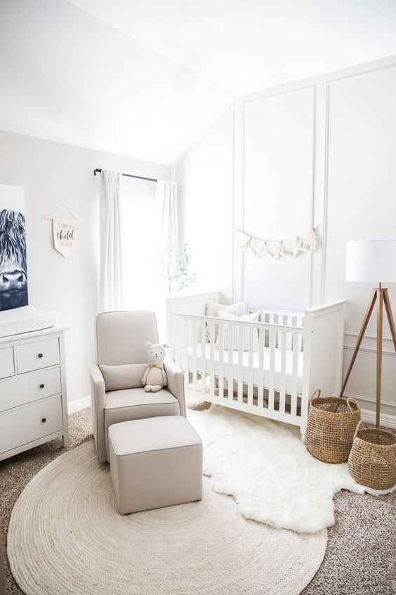 a small white nursery with paneled walls, a white crib, a white dresser, layered rugs, a gray chair with an ottoman and some decor