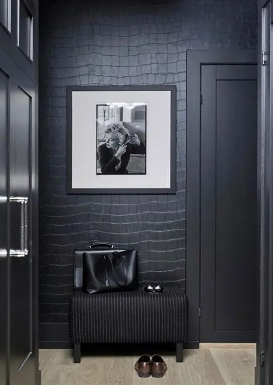 A soot-colored entryway with reptile-patterned wallpaper, a black stool, a piece of art and a few pieces of furniture is very chic and sophisticated