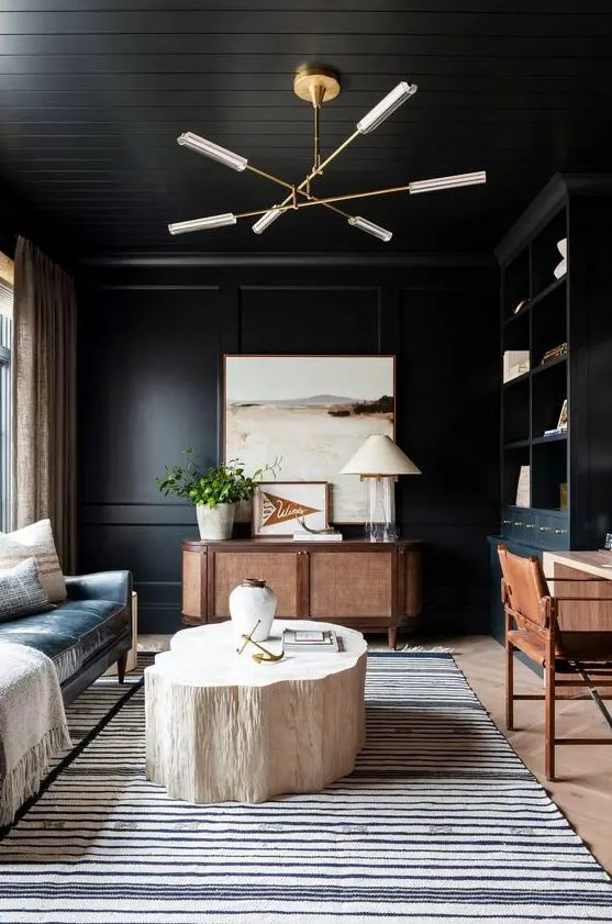 a sooty home office with a black leather sofa, a stained credenza, a desk and chair, a chandelier and a bleached stump table