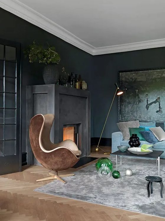 a sooty living room with a fireplace, dark artwork, a blue sofa and colorful cushions, a brown egg-shaped chair and some vases