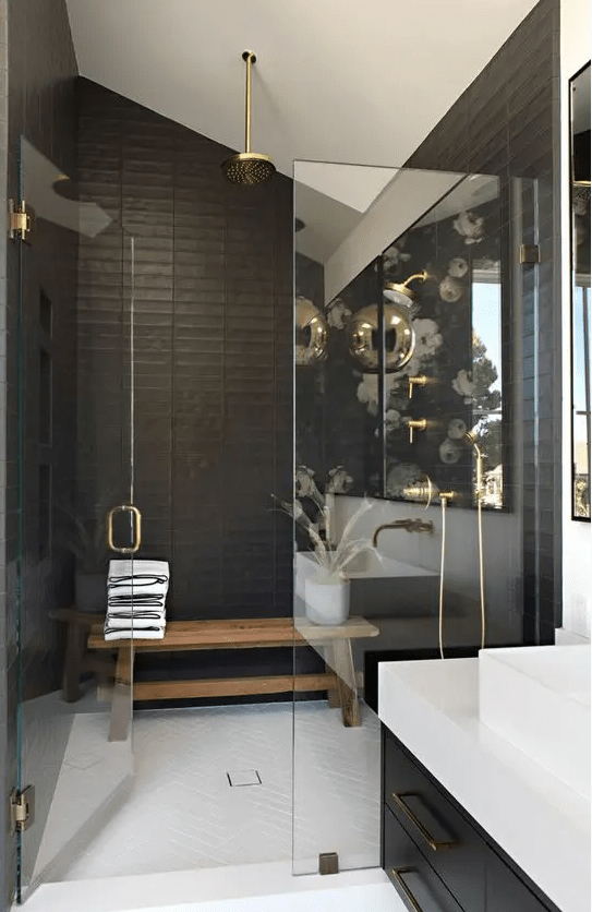 a stylish black and white bathroom with a shower area, a bench, gold fixtures, a black vanity and a white stone countertop