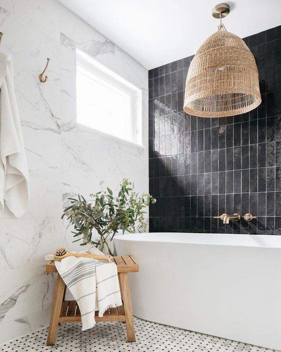 a stylish black and white bathroom with marble and black stacked tiles, a bathtub, a woven pendant lamp and a stained stool