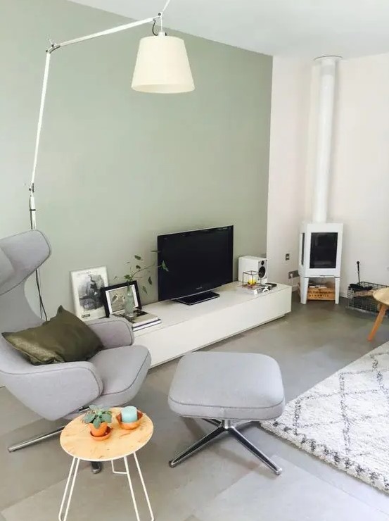 a stylish living room with a light green accent wall, a television set and a television, a white fireplace, a gray chair with an ottoman, some decor and a lamp