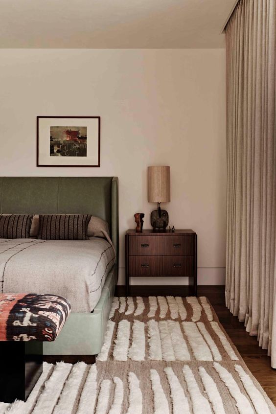 a subtly earthy bedroom with gray curtains and a taupe rug, a green bed with neutral linens, nightstands and a statement bench