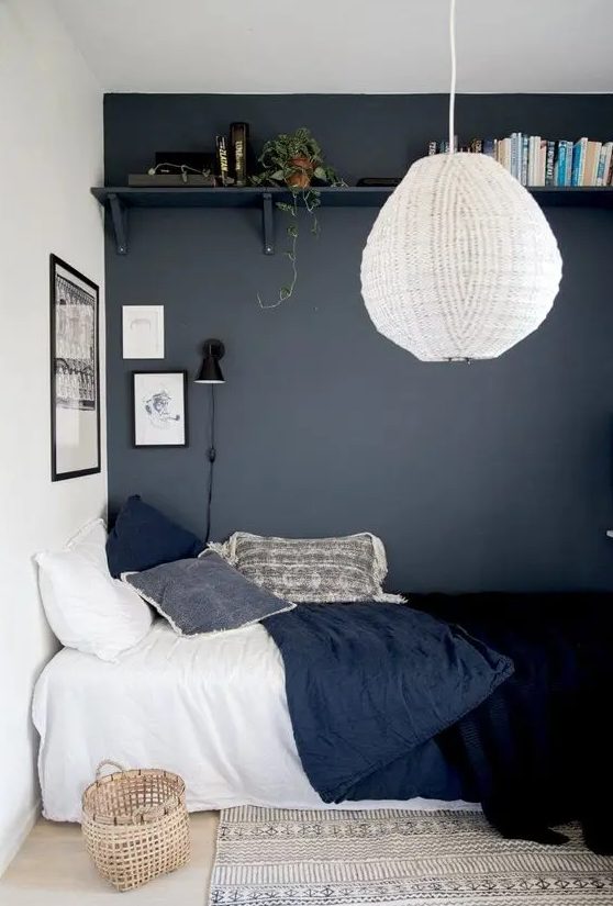 a teenager's room with a graphite gray wall, an open shelf, a bed with dark blue linens, a basket, a pendant lamp and a carpet