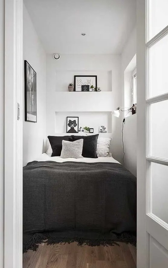 A tiny bedroom in white, with just a squashed bed, black and white bedding, niche shelves with decor, a piece of art and a sconce