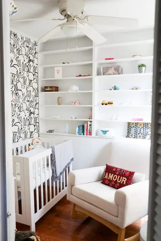 A small, neutral nursery with a bunny accent wall, built-in shelves, a white crib and rocker, and some bright textiles