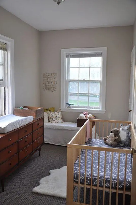 A small, neutral nursery with a daybed, a mid-century modern dresser that doubles as a changing table, a crib with neutral bedding, and layered rugs is amazing