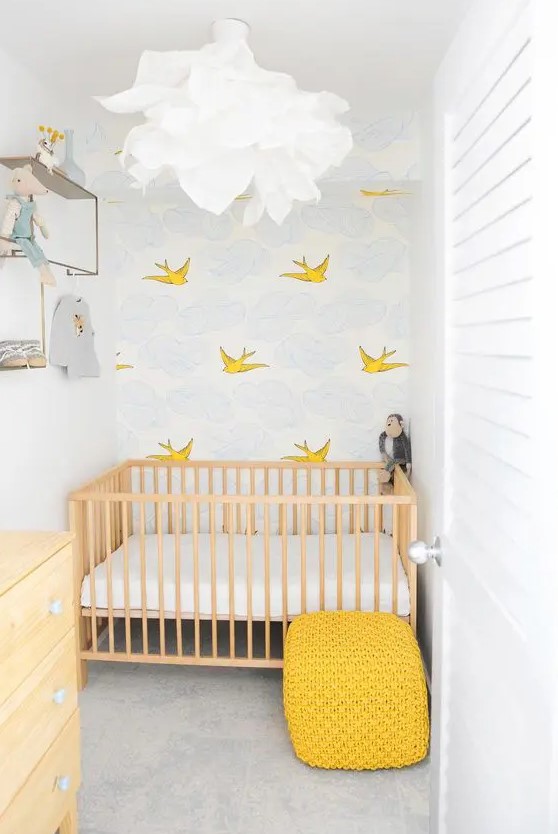 a small nursery with an accent wall, a stained crib with neutral linens, a stained dresser, a yellow stool and some shelves