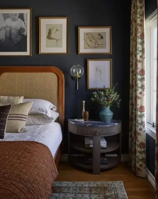 a moody vintage bedroom with sooty walls, a bed with a cane headboard, a fluted nightstand, a gallery wall and printed textiles