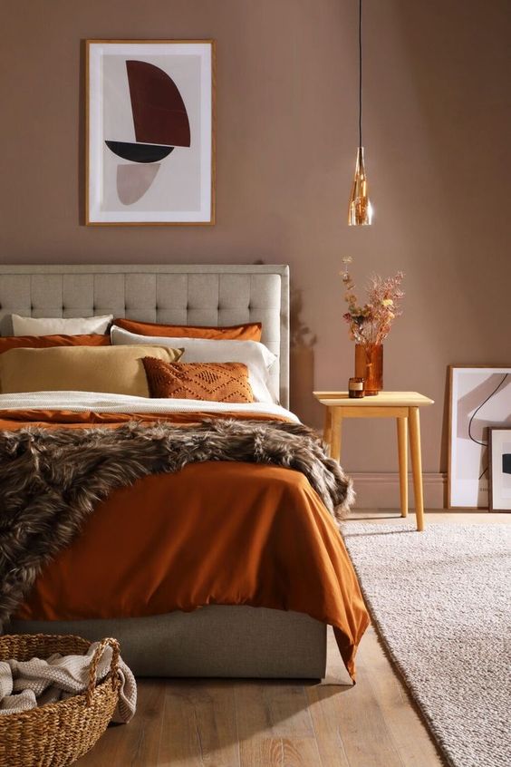 a bedroom decorated in warm earth tones with a taupe accent wall, a gray bed with light linens, a stool with dried herbs and a pendant lamp