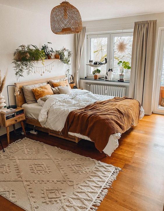 An inviting, earthy boho bedroom features stained furniture, rust and white linens, a patterned rug, lots of potted plants, and a woven hanging lamp