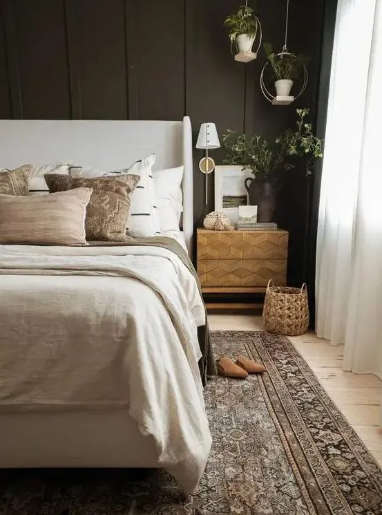 An inviting, moody bedroom with a soot-colored accent wall, a neutral upholstered bed with neutral linens, a printed rug, hanging plants and an inlaid nightstand