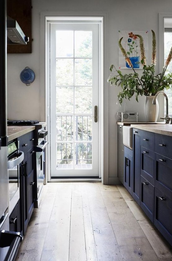 An inviting navy blue farmhouse kitchen with navy blue cabinets and wood countertops, a hardwood floor and lots of natural light through the door