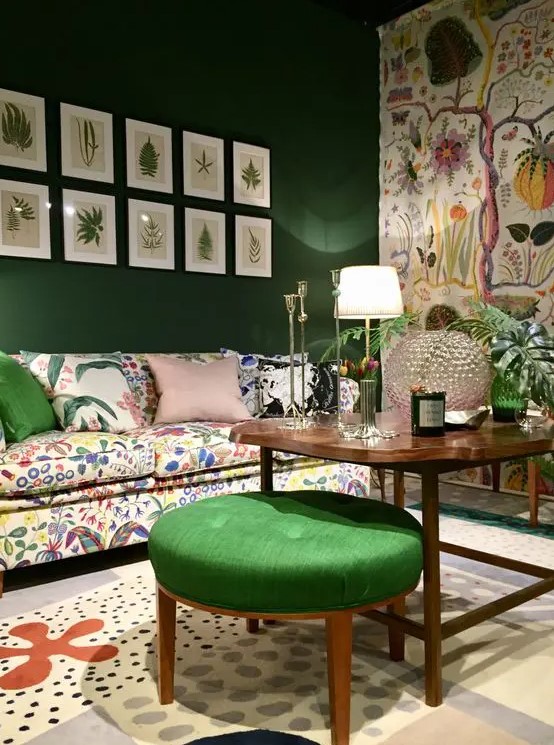 a whimsical living room with an emerald green and a floral accent wall, a crazy floral sofa and rug, a gallery wall and a green stool