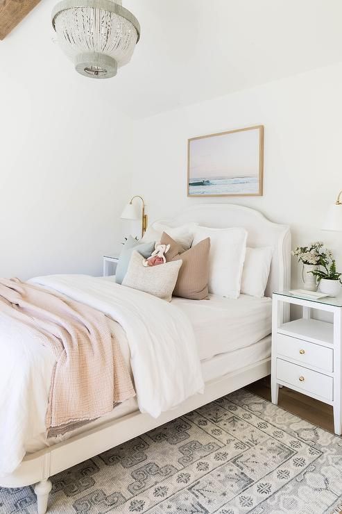 a white French double bed with a pale pink blanket, a white bedside table and a white and gray carpet, lit by a white and gray beaded chandelier