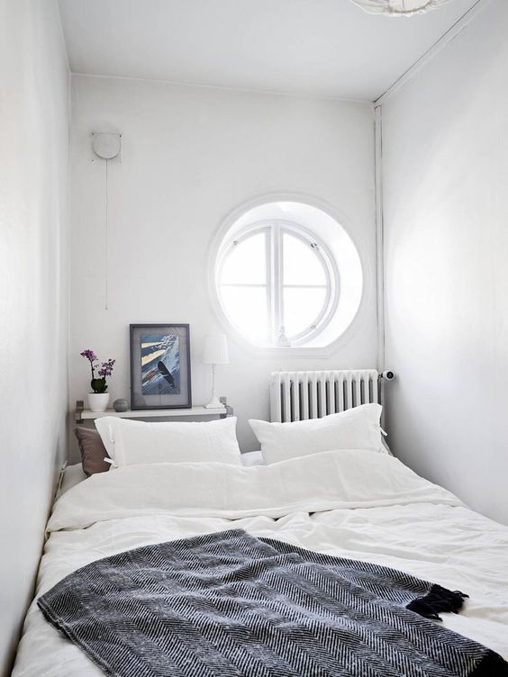 a white, narrow bedroom with a porthole, a bed with neutral linens, a narrow bedside table at the headboard and a radiator