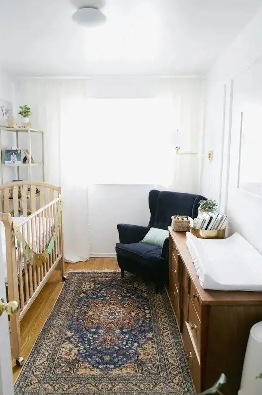 A white nursery features a navy velvet chair, a printed rug and a brown wooden dresser
