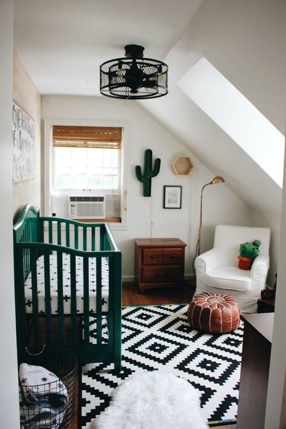 A small attic children's room with a printed rug, a green crib, a white chair, stained furniture, decor and a black hanging lamp