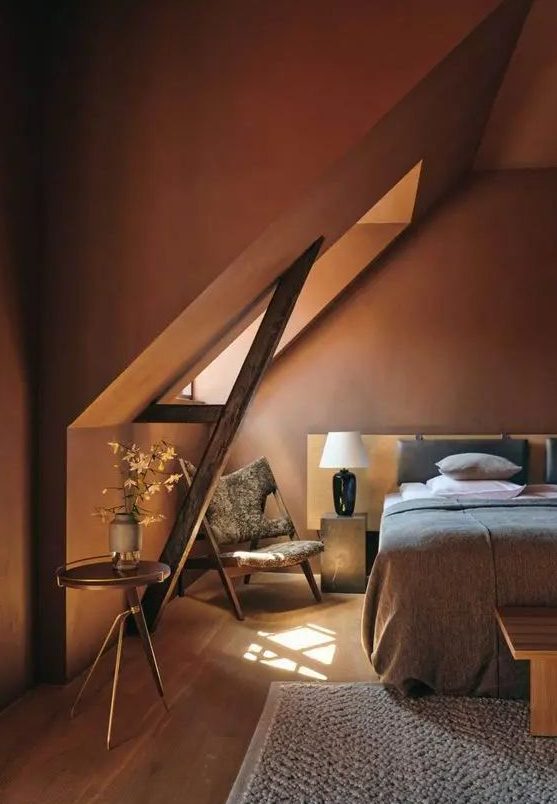 an earthy attic bedroom with burgundy walls, a bed with neutral linens, stone nightstands and a statement chair