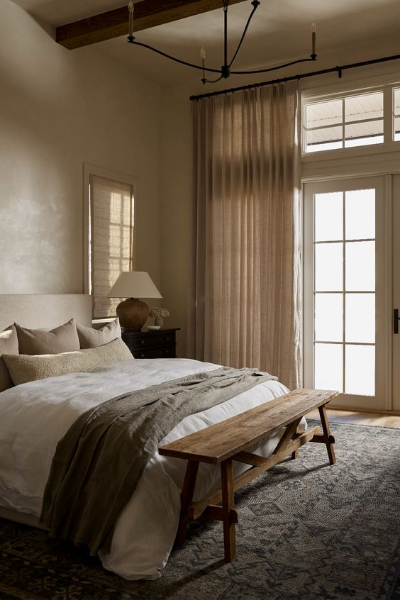 an earthy bedroom with light brown walls, a modern neutral bed with neutral linens, a wooden bench and neutral textiles