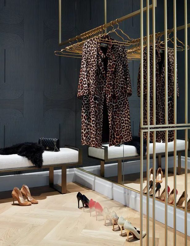 an elegant closet with a soot patterned wall, a mirrored wall, a neutral bench, gold railings and hangers