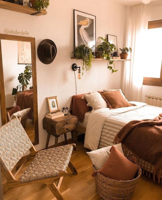 An inviting earth-toned bedroom features a bed with rust and neutral linens, a basket of pillows, a wicker chair and some potted plants