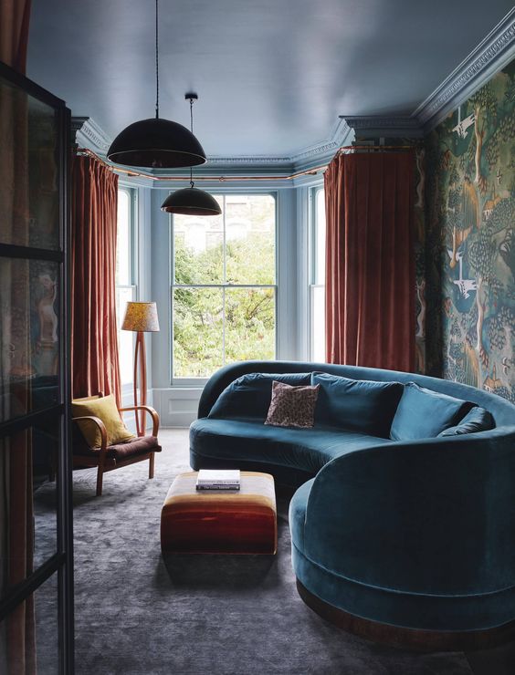 A moody and bold living room with a curved blue sofa, orange textiles and moody bird pattern wallpaper on one wall