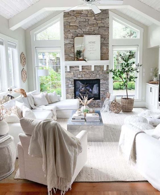 A white farmhouse living room with a stone-clad fireplace, white furniture, potted plants, neutral fabrics and a view
