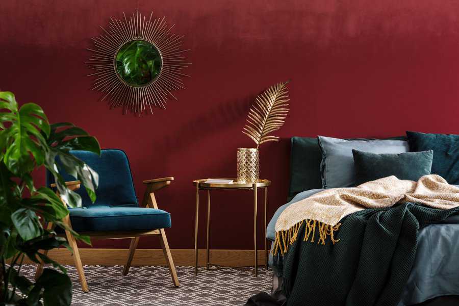 Gold table between green armchair and bed in elegant red bedroom interior with mirror