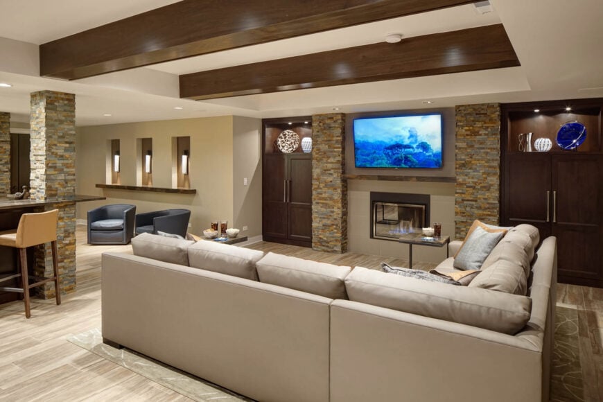 The thoroughly modern family room combines an open plan with the kitchen hinted at left, light hardwood floors and a series of stone columns under a white ceiling with exposed, polished wooden beams. A sharply defined L-shaped sectional dominates the center, while rich wooden furniture flanks a fireplace and television area.