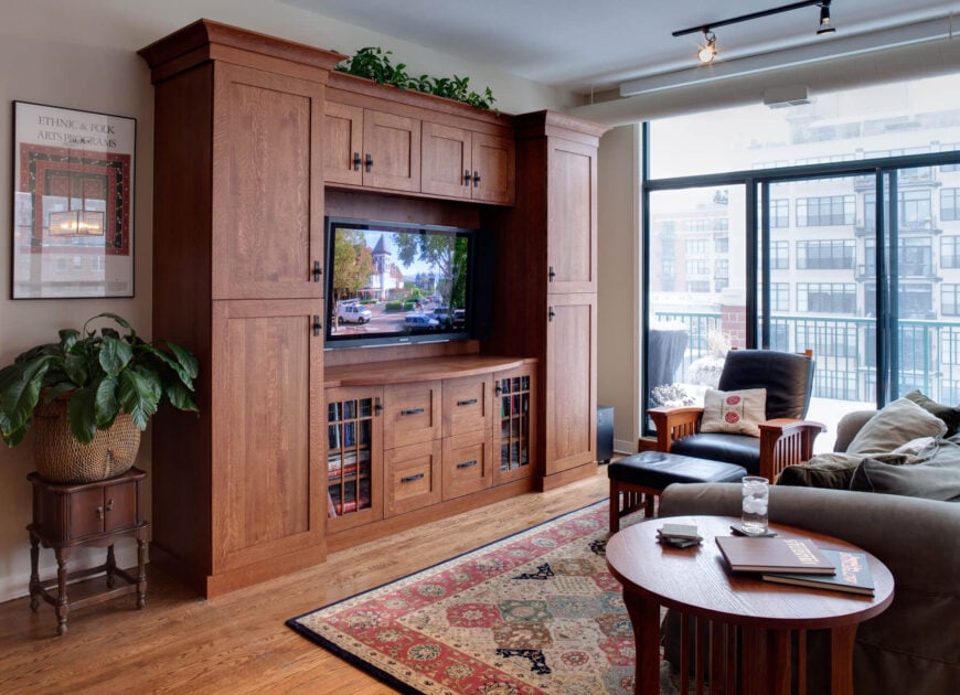 This high-rise family room exudes a natural setting with hardwood floors and warm floor-to-ceiling wood cabinets housing the center TV. The wooden-framed armchair with leather cushions sits in front of floor-to-ceiling sliding glass doors, allowing expansive city views and access to the balcony.