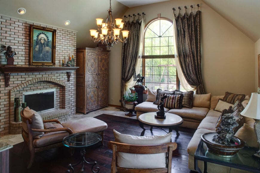 Bespoke luxury defines this cozy family room, framing rich hardwood floors with tile all around. A large brick fireplace with a carved wooden mantle commands attention, while in the center hangs an old-fashioned chandelier that peaks through an arched, floor-to-ceiling window. The L-shaped section balances a pair of wooden-framed chairs and round coffee tables.