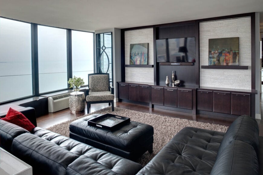 A sumptuous button-tufted black leather sofa envelops this family room, showcasing hardwood floors, wall-length dark wood shelves, and a brown shag rug. Floor-to-ceiling windows illuminate the space.