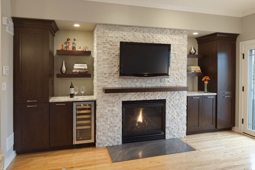 A sharp contrast defines this living room with dark wood cabinets flanking a white textured brick fireplace surround on light, natural hardwood flooring. The fireplace faces a slab of dark marble, while the cabinets feature a built-in wine cooler and marble countertops beneath floating wooden shelves.
