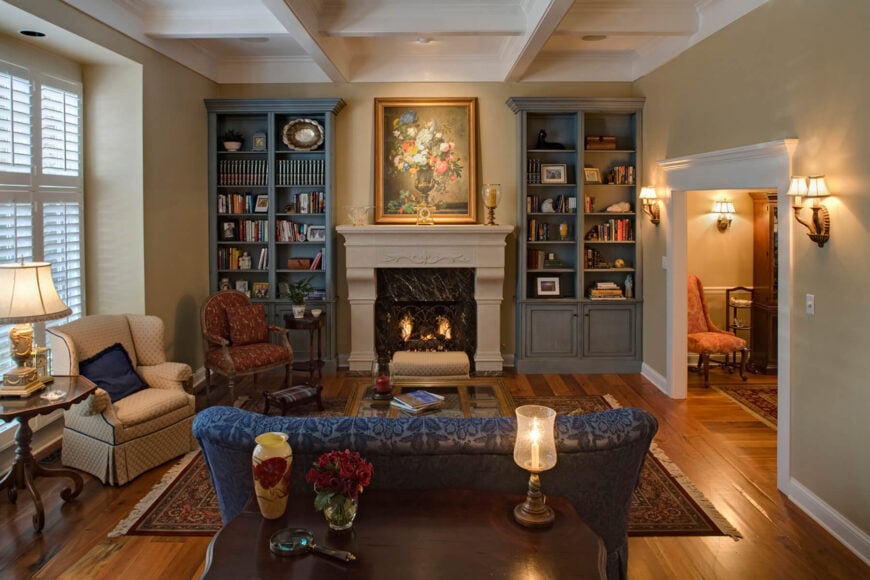 Lush details and a variety of tones abound in this beautiful living room, with a detailed white ceiling over beige walls and rich hardwood flooring. A pair of full-height gray bookcases flank the central stone fireplace, while a row of chairs in blue, brown, and red, respectively, surround a patterned rug with a mirror-top coffee table.