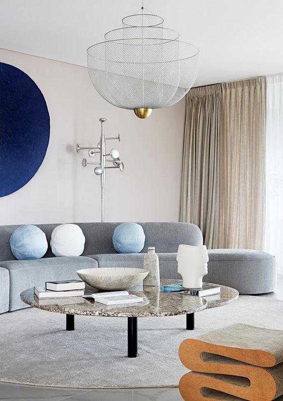 A neutral, statement living room with a curved gray sofa, navy blue artwork, and a marble coffee table and cork stool