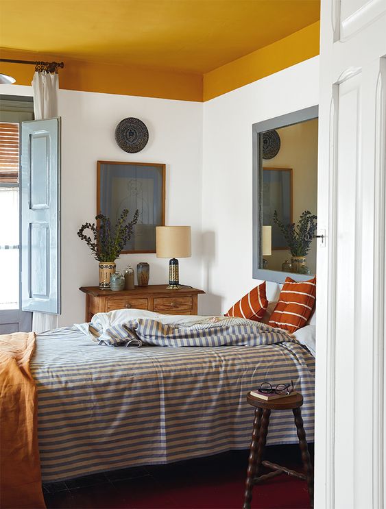 a Mediterranean bedroom with a mustard yellow blanket, a bed with printed linens, a stained console table and a large mirror