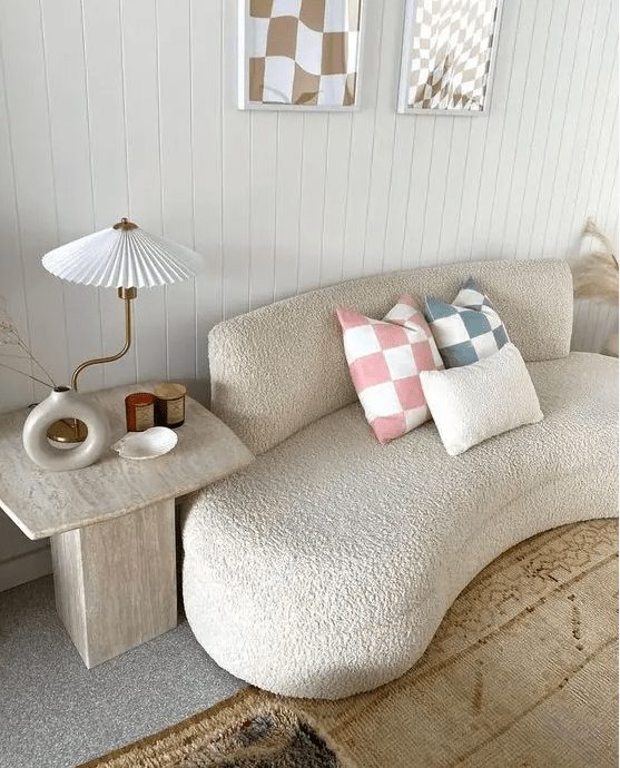 a curved bouclé sofa with printed cushions, a stone side table, some candles, a vase and a whimsical lamp