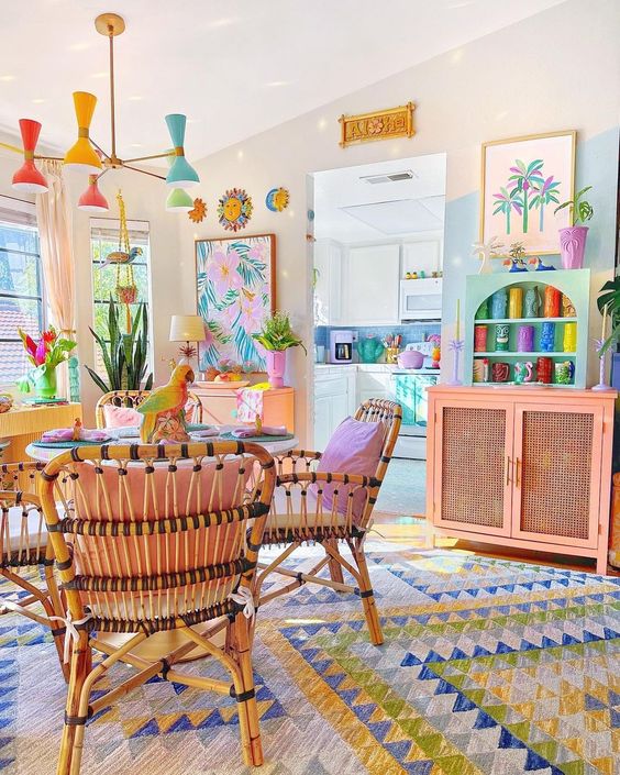A bright living room with dopamine decor, a table and rattan chairs, a statement rug, a pink cabinet and turquoise shelves, and a pastel chandelier