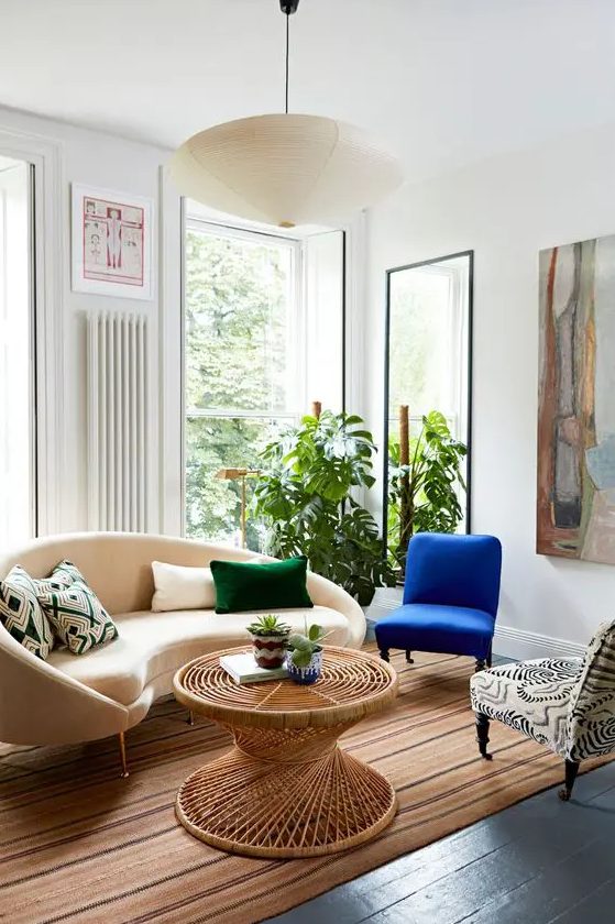 a bright living room with a curved sofa and cushions, a blue and a printed chair, a rattan coffee table, some art and a mirror