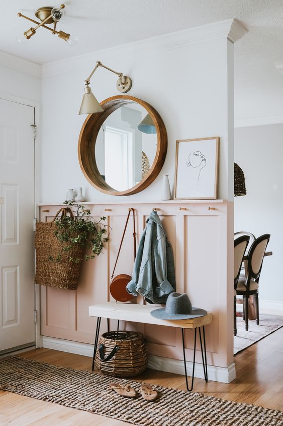 A chic, modern entryway with a pink paneled wall, a round mirror, a bench with pinhair legs and baskets