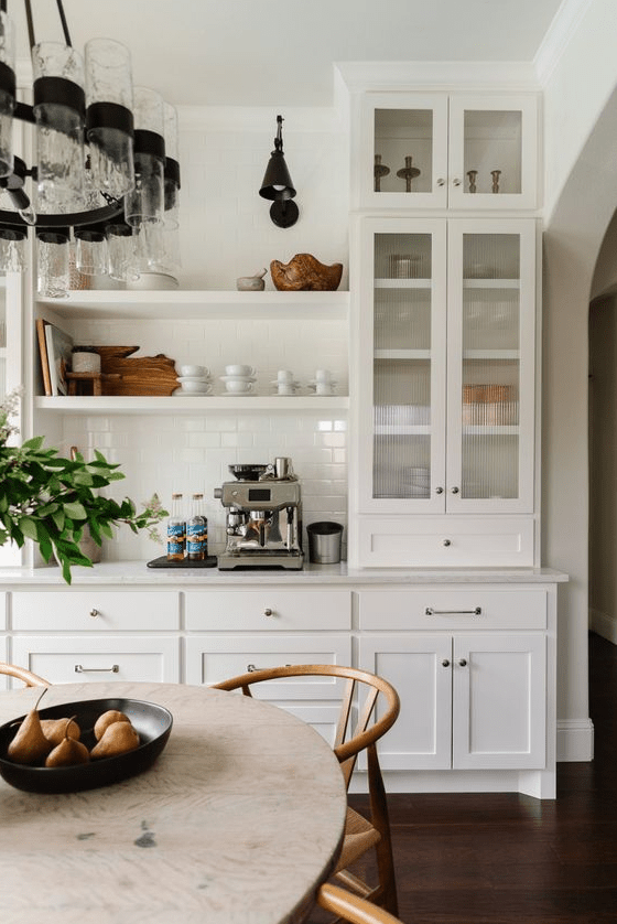a chic white shaker-style kitchen with glass-front cabinets, built-in shelving, a white tile backsplash and white marble countertops