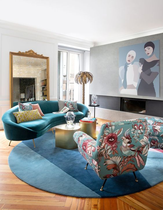 a colorful and eclectic living room with a built-in fireplace, a large piece of art, a blue curved sofa and rug, a floral chair and gold coffee tables