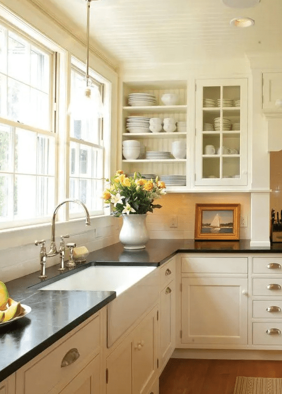 a cream vintage kitchen with shaker and glass cabinets, black quartz countertops, a window backsplash, and knobs and pulls