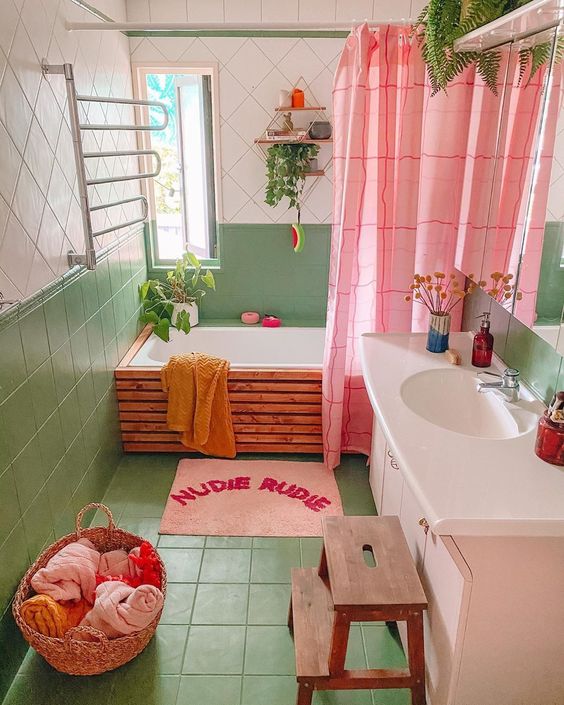 a bathroom in dopamine decor with green and white tiles, a wooden bathtub, a blush vanity, a pink curtain and a rug